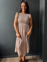 Woman wearing a beige dress standing against a dark wall.
