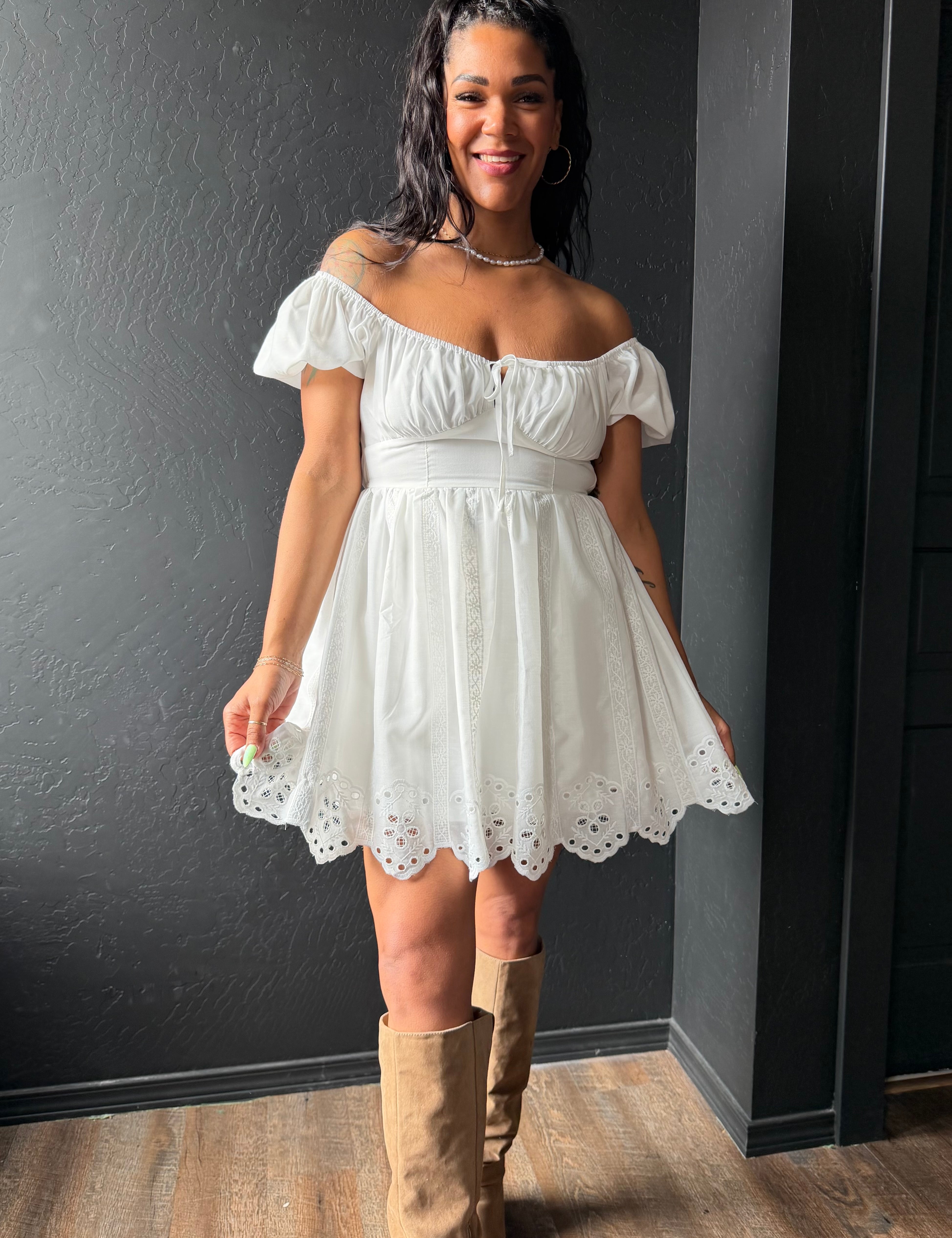 Woman wearing a white off-shoulder dress with lace details and brown knee-high boots against a dark wall.