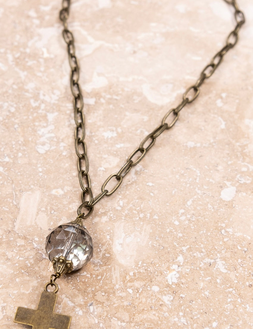 A bronze-colored cross pendant attached to a chain link necklace, featuring a clear crystal pendant below the cross.