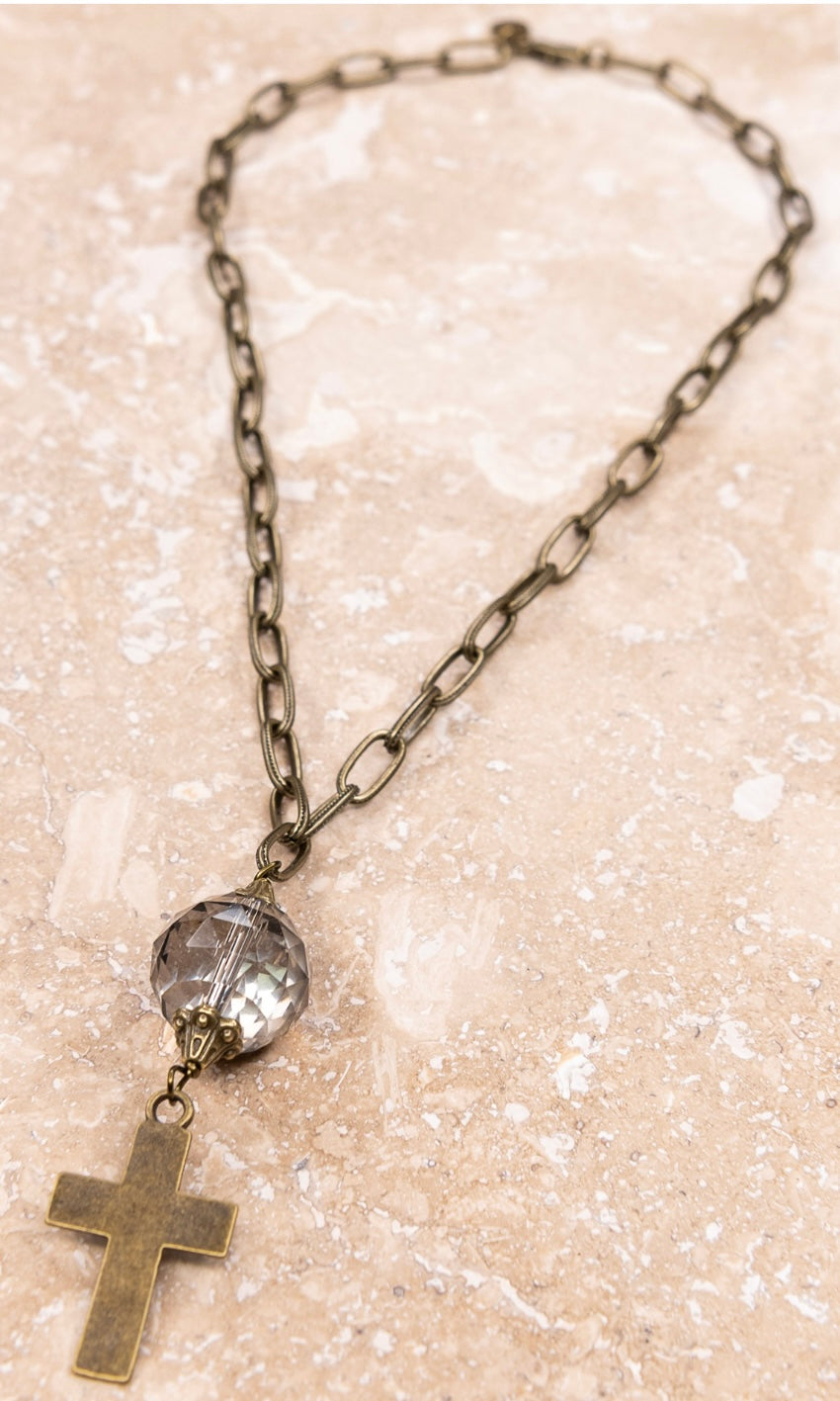A bronze-colored cross pendant attached to a chain link necklace, featuring a clear crystal pendant below the cross.