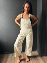 Woman wearing beige overalls standing against a dark wall.
