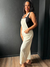 Woman wearing white overalls and a black top against a dark wall.