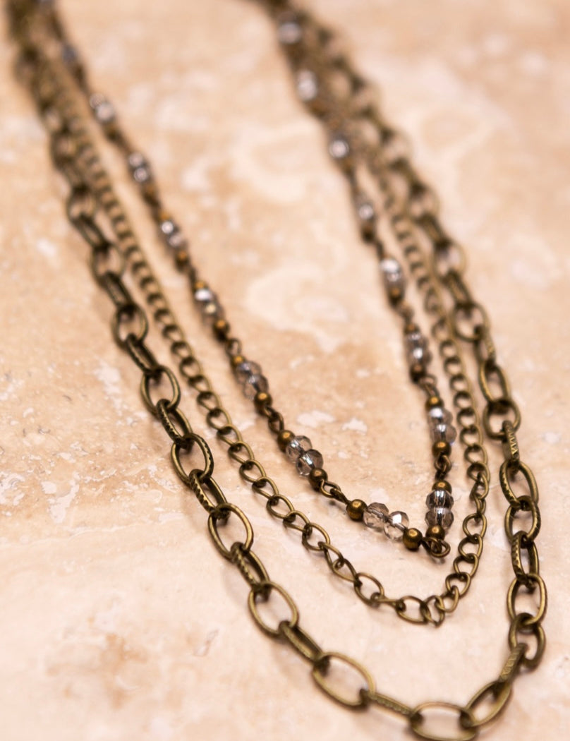 A multi-layered bronze beaded necklace with a chain link strand, laid out on a textured surface.