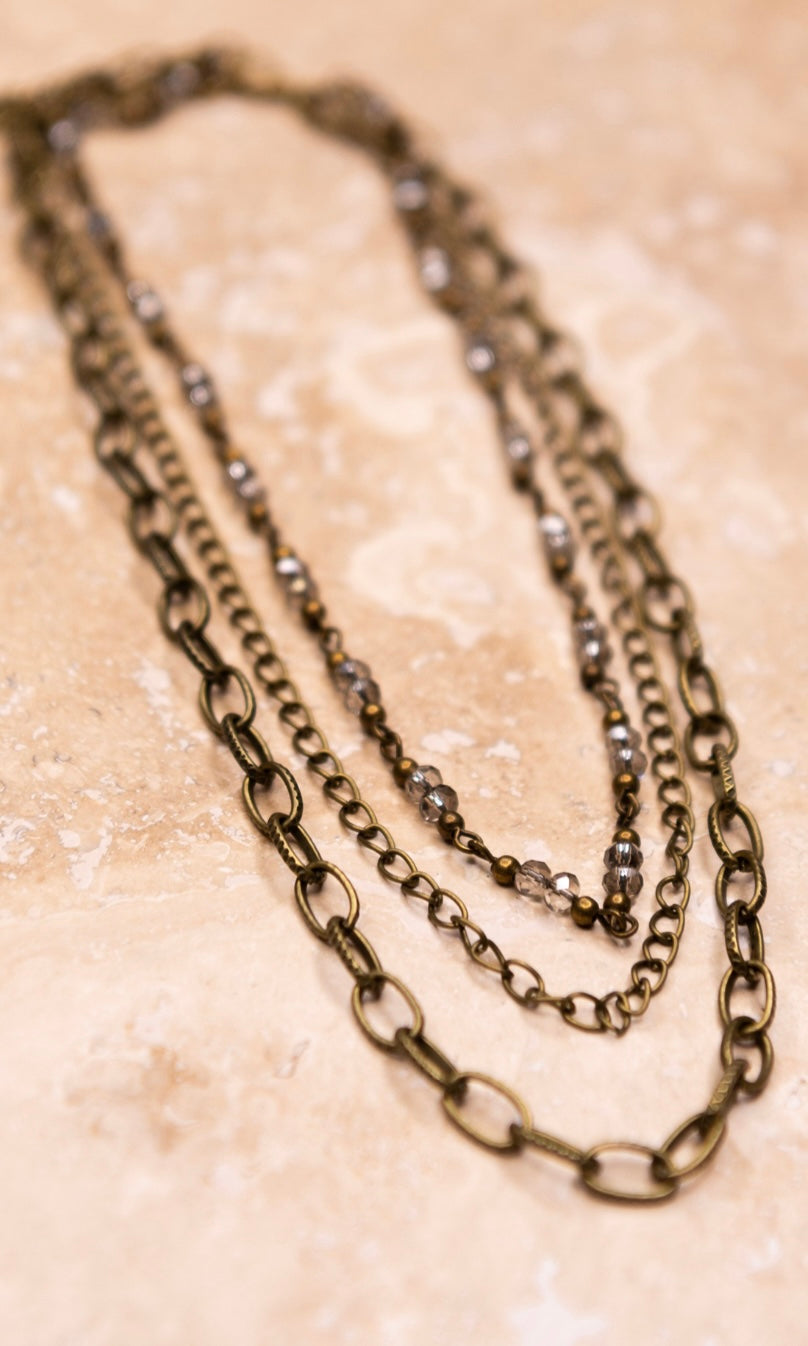 A multi-layered bronze beaded necklace with a chain link strand, laid out on a textured surface.