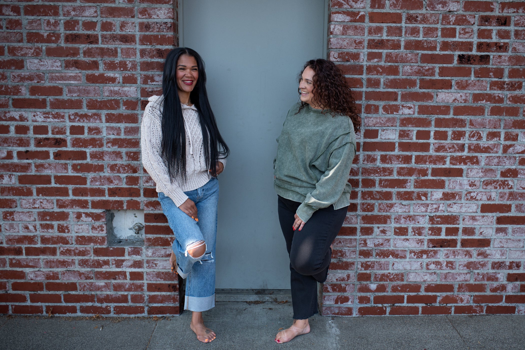 Two women standing barefoot in front of a red brick wall and gray door, wearing casual sweatshirts and jeans.