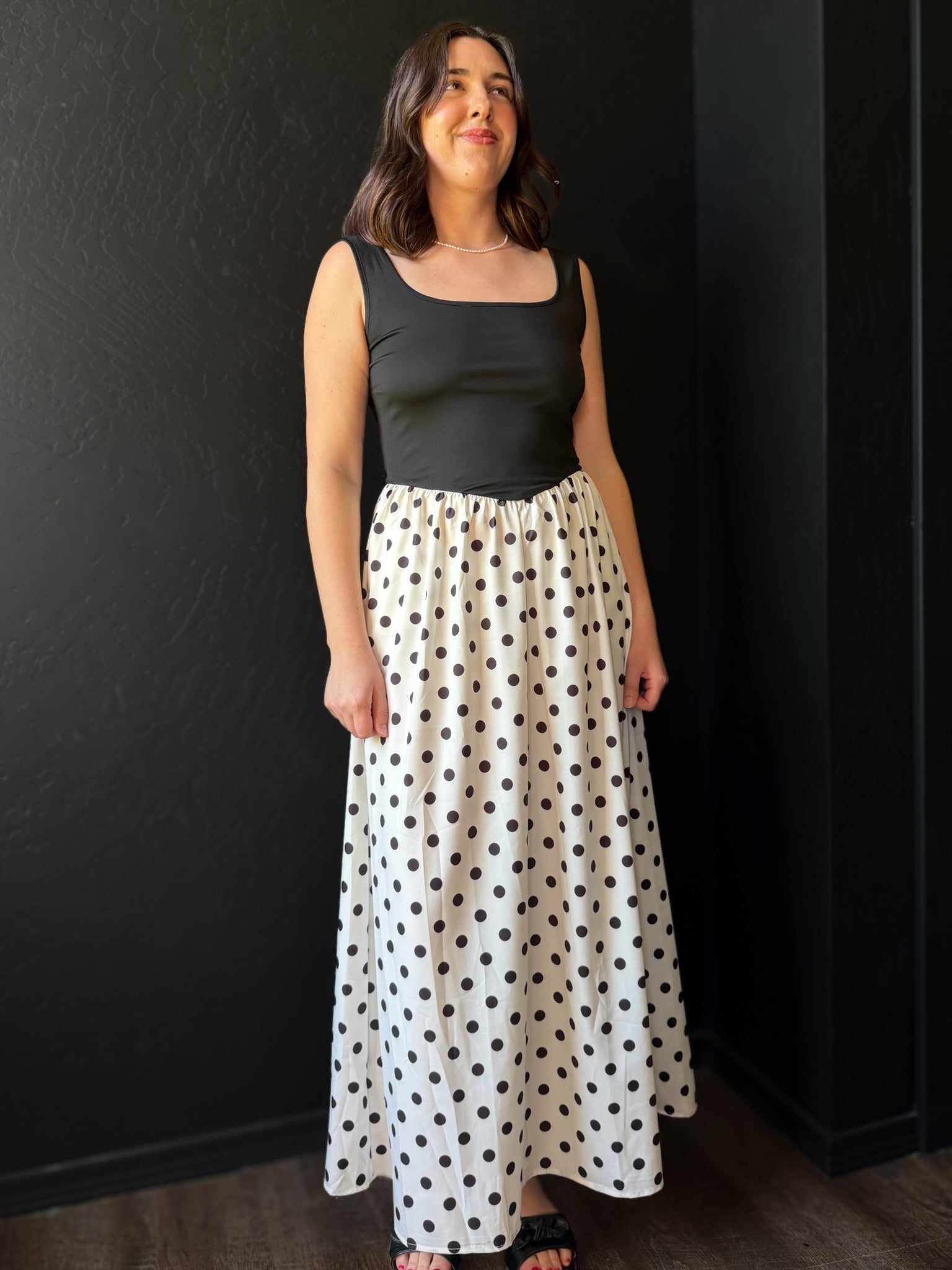 Woman wearing a black tank top and polka dot skirt against a dark wall.