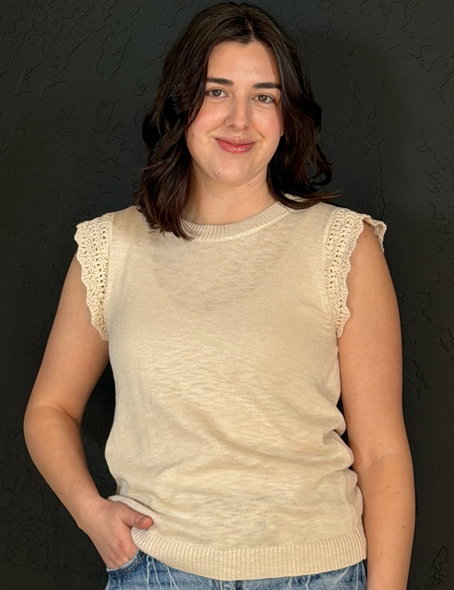 Woman wearing a beige sleeveless sweater with lace trim against a dark background