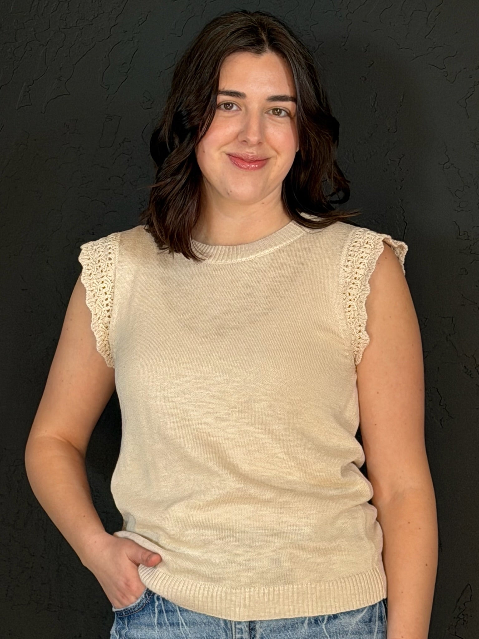 Woman wearing a beige sleeveless sweater with lace trim against a dark background