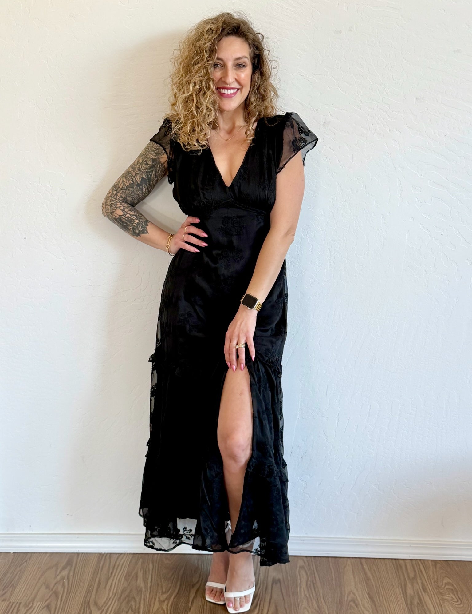 Woman wearing a black dress with a slit standing against a white wall.