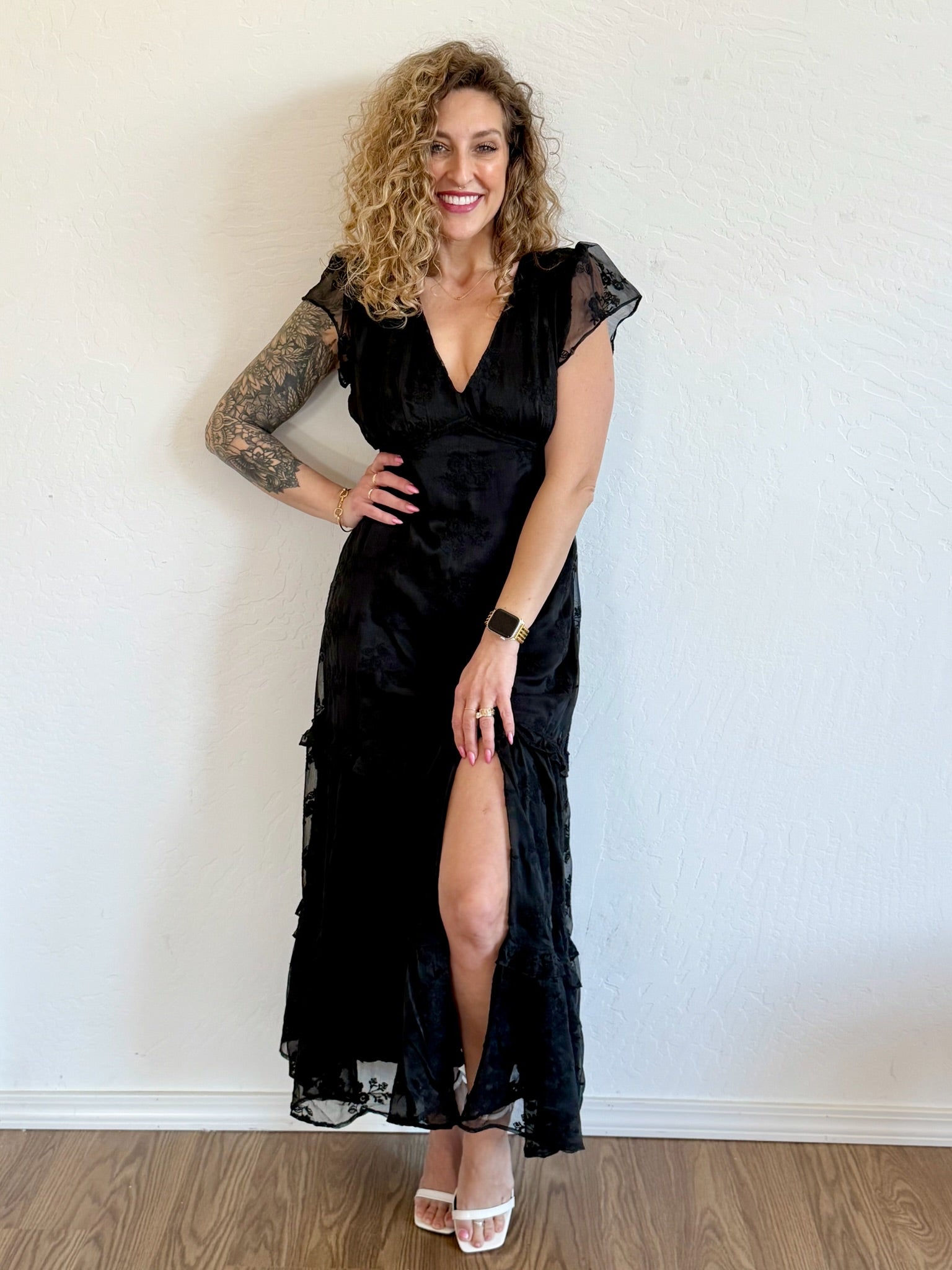 Woman wearing a black dress with a slit standing against a white wall.