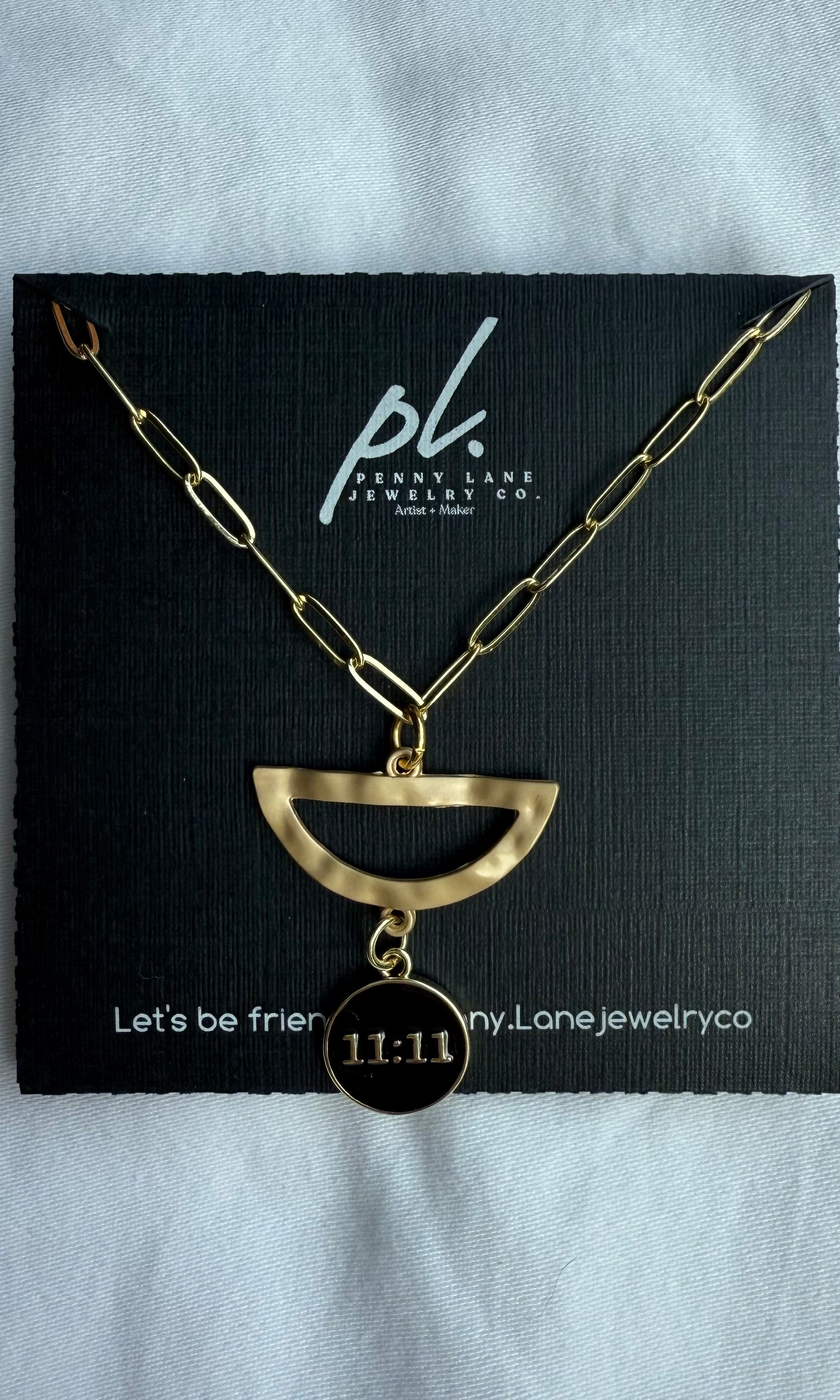 Gold necklace with a half-circle pendant and '11:11' charm on a black card with 'penny lane jewelry co' branding.