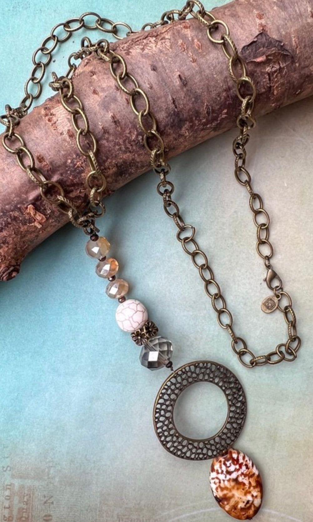 A long necklace with a round, textured metal pendant and a variety of natural polished stones strung along the bottom half. The pendant is gold in color, and the stones vary in hue, set on a chain link.