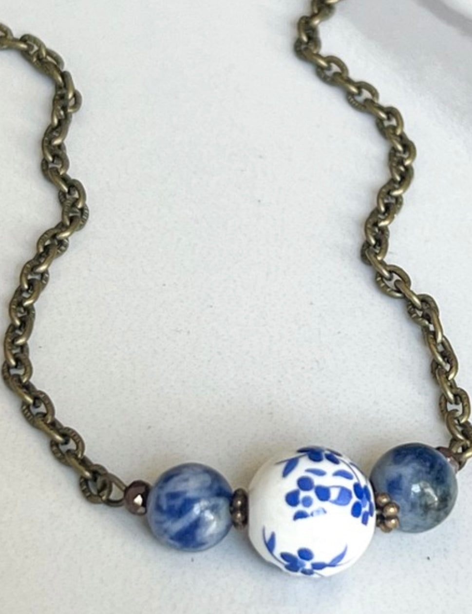 A bronze chain necklace with three blue and white floral beads in the center displayed against a marble background.