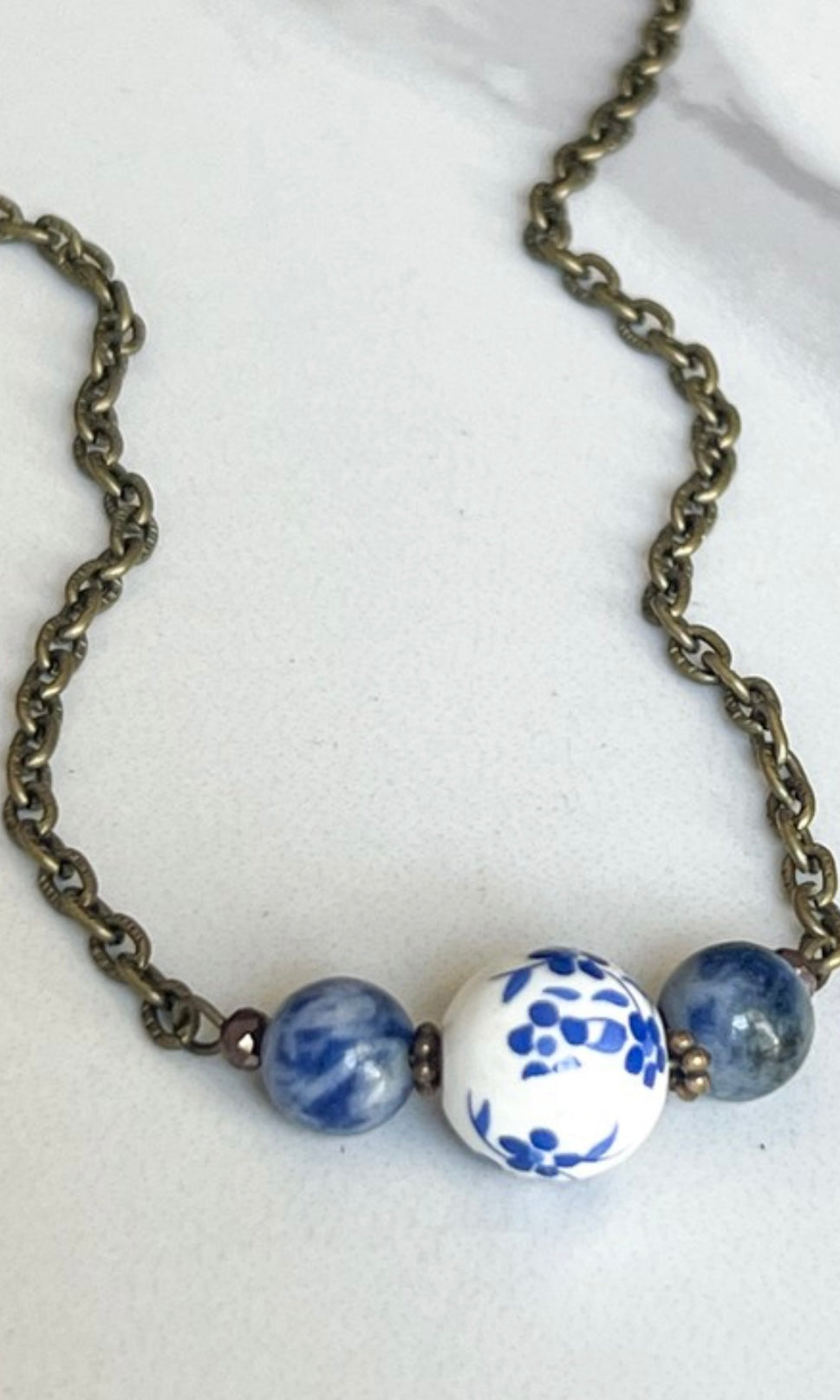 A bronze chain necklace with three blue and white floral beads in the center displayed against a marble background.