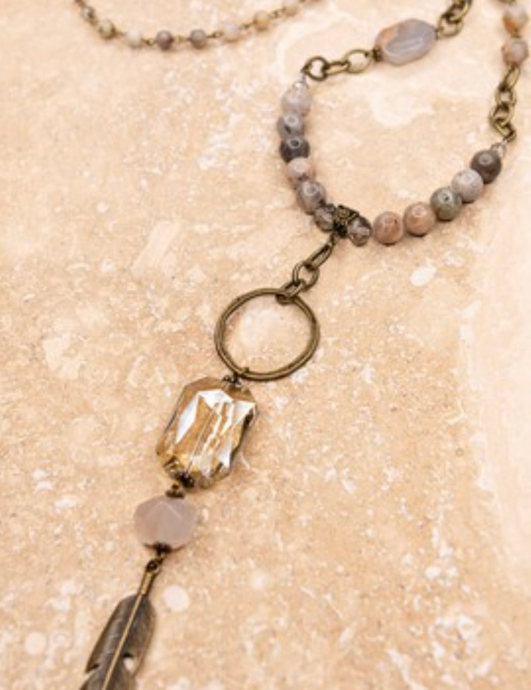 A long beaded necklace with a round drop pendant featuring a crystal stone and a feather design, ending with an adjustable lobster claw closure.