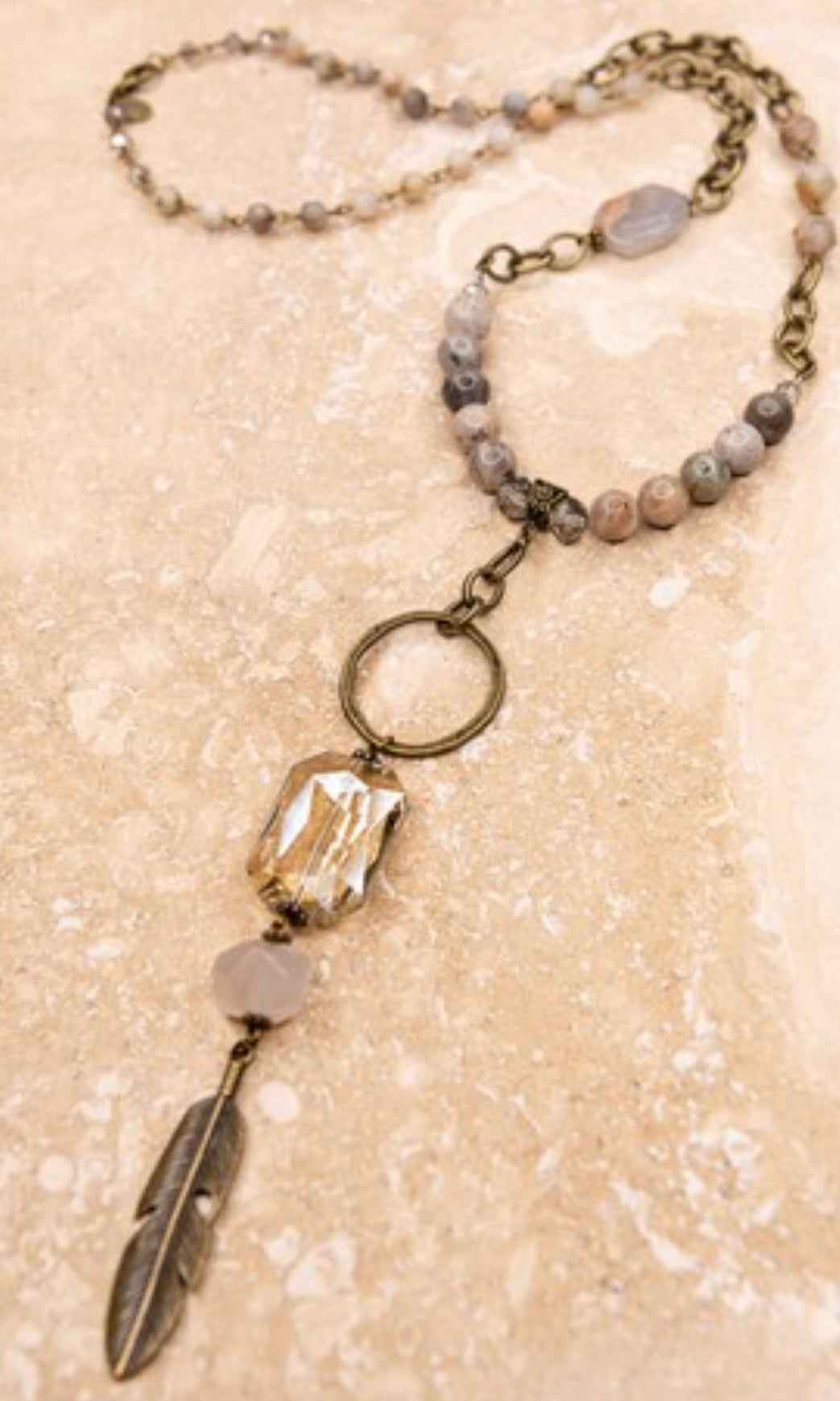 A long beaded necklace with a round drop pendant featuring a crystal stone and a feather design, ending with an adjustable lobster claw closure.