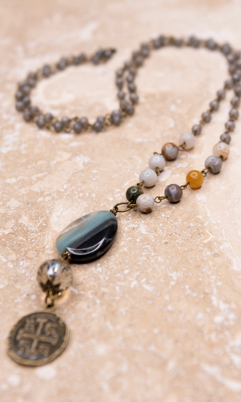 A long beaded chain link necklace with a grey stone pendant and a metal doubloon coin pendant.