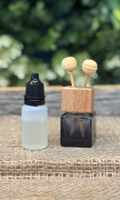 Small glass bottle with black cap, wooden holder with two wooden sticks, and black square container on a textured surface with blurred greenery in the background.