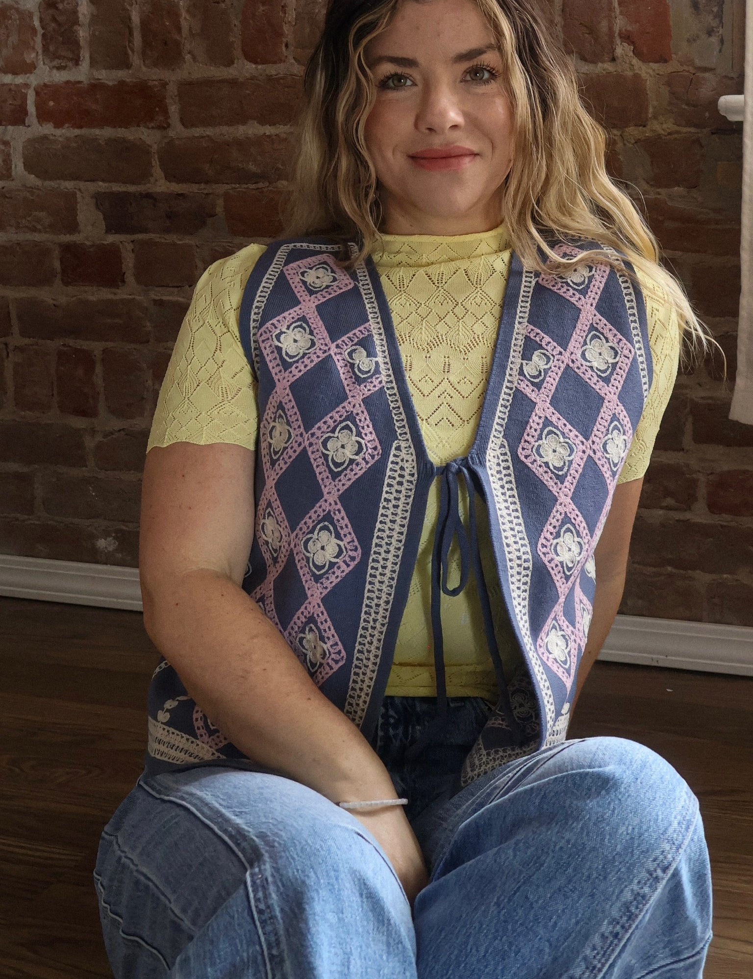 Woman wearing a patterned vest sitting against a brick wall.