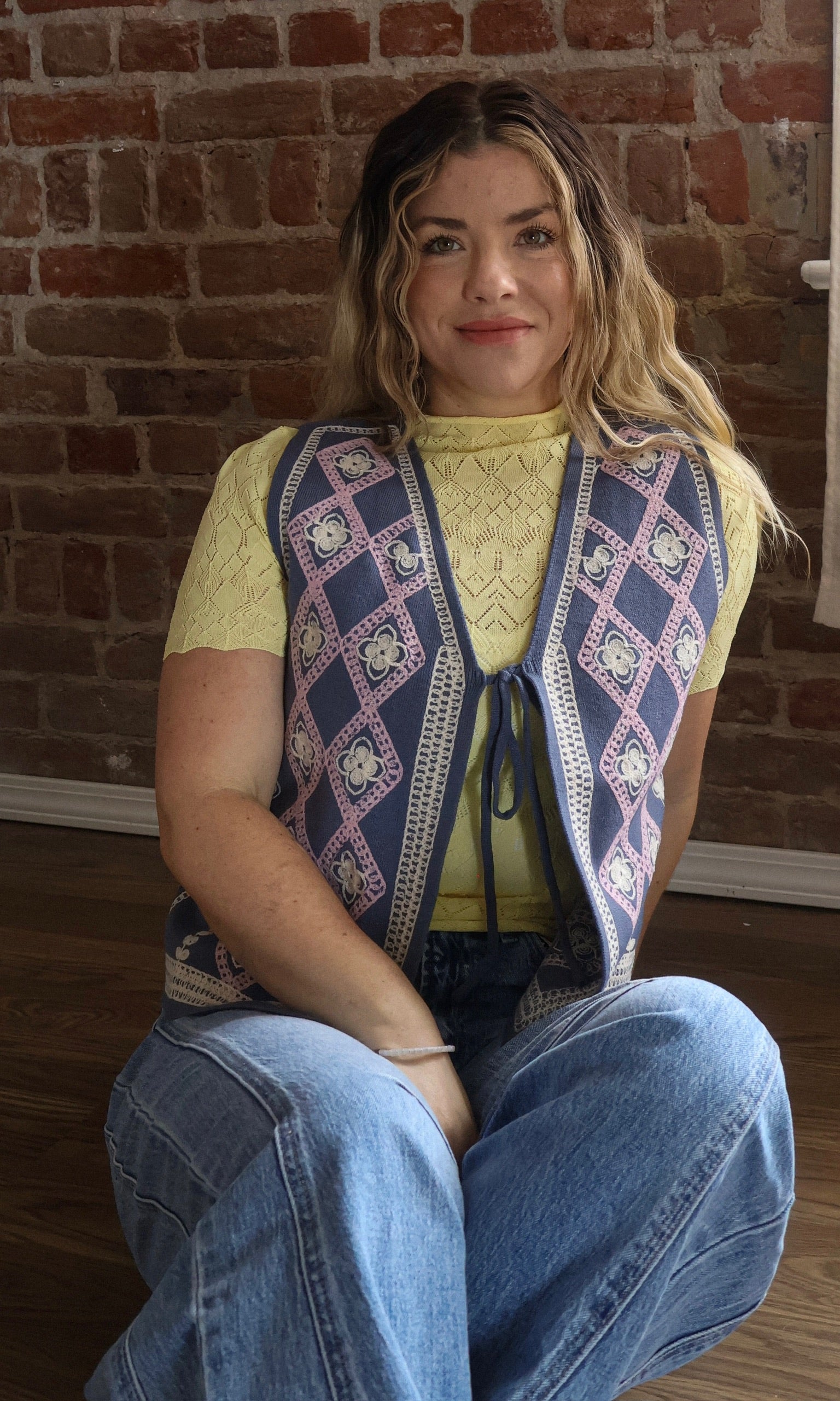 Woman wearing a patterned vest sitting against a brick wall.