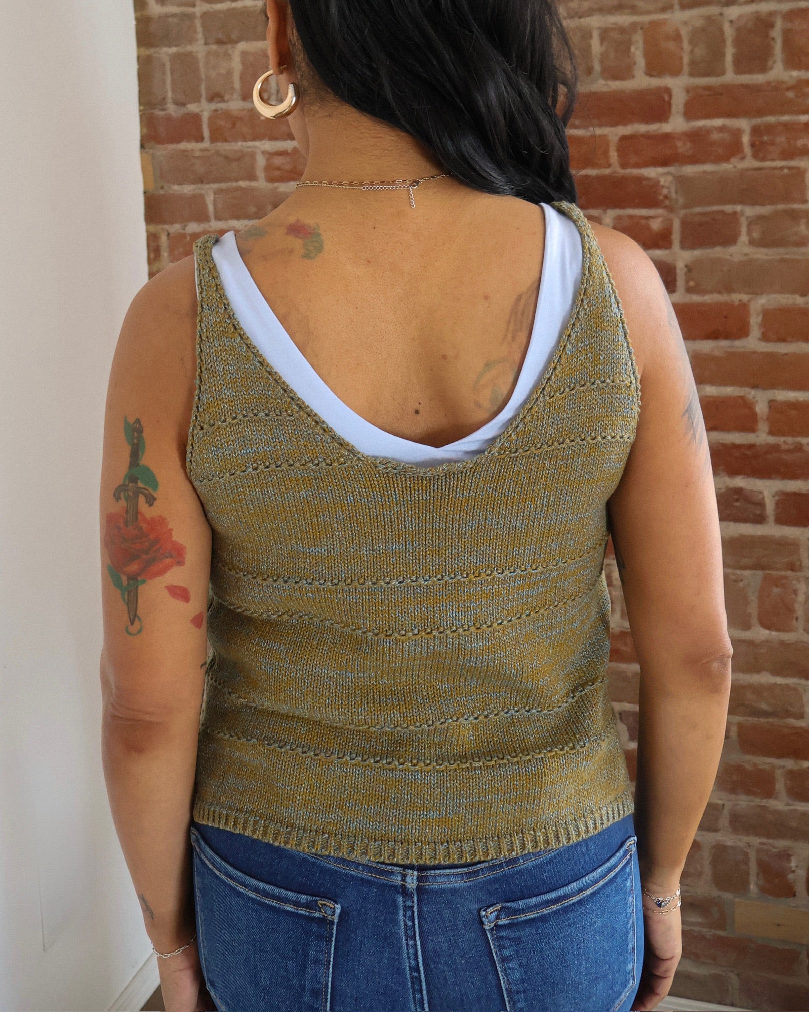 Person wearing a green knitted tank top with a white bralette underneath, standing against a brick wall.