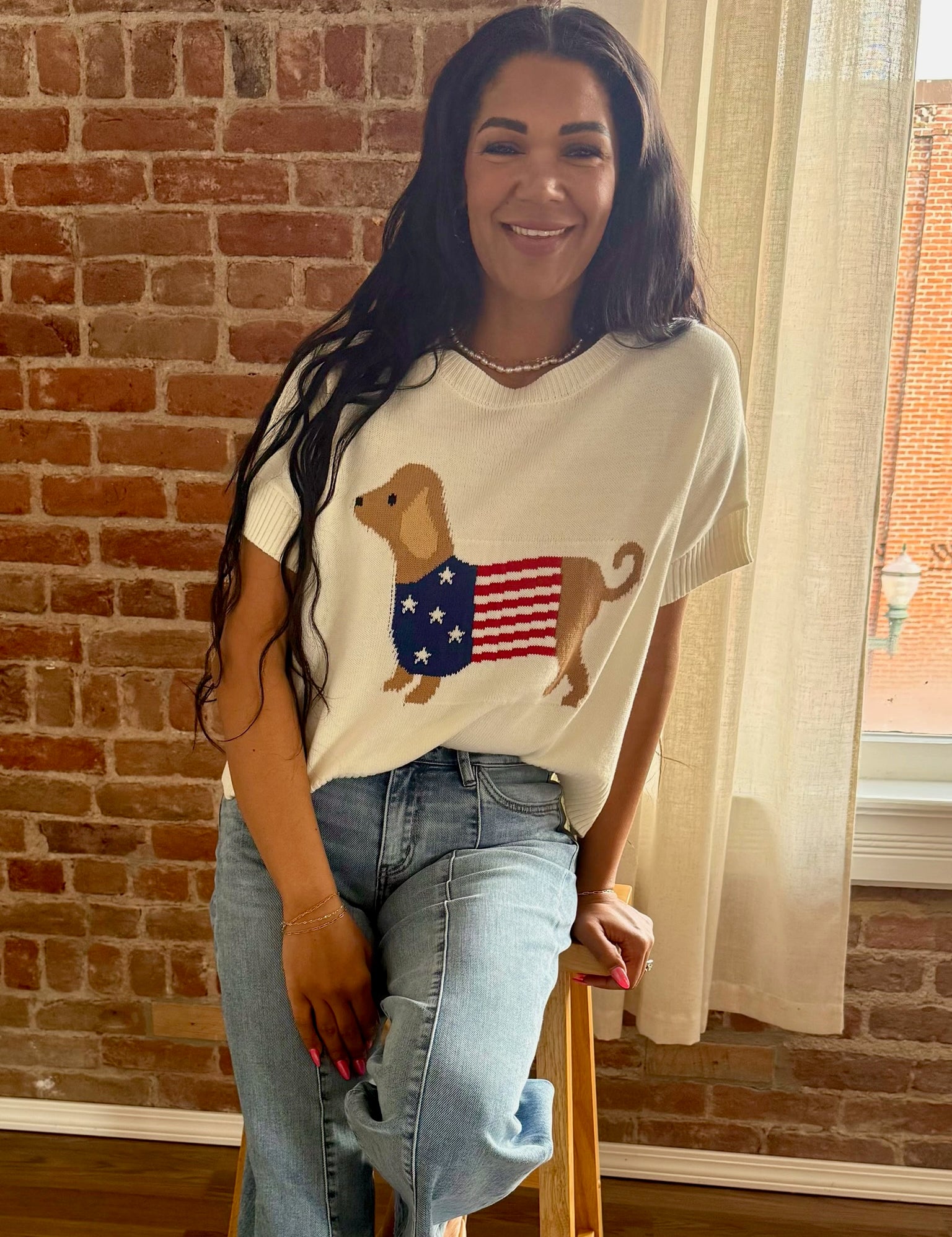 Woman wearing a white t-shirt with a dog and American flag design, standing in front of a brick wall.