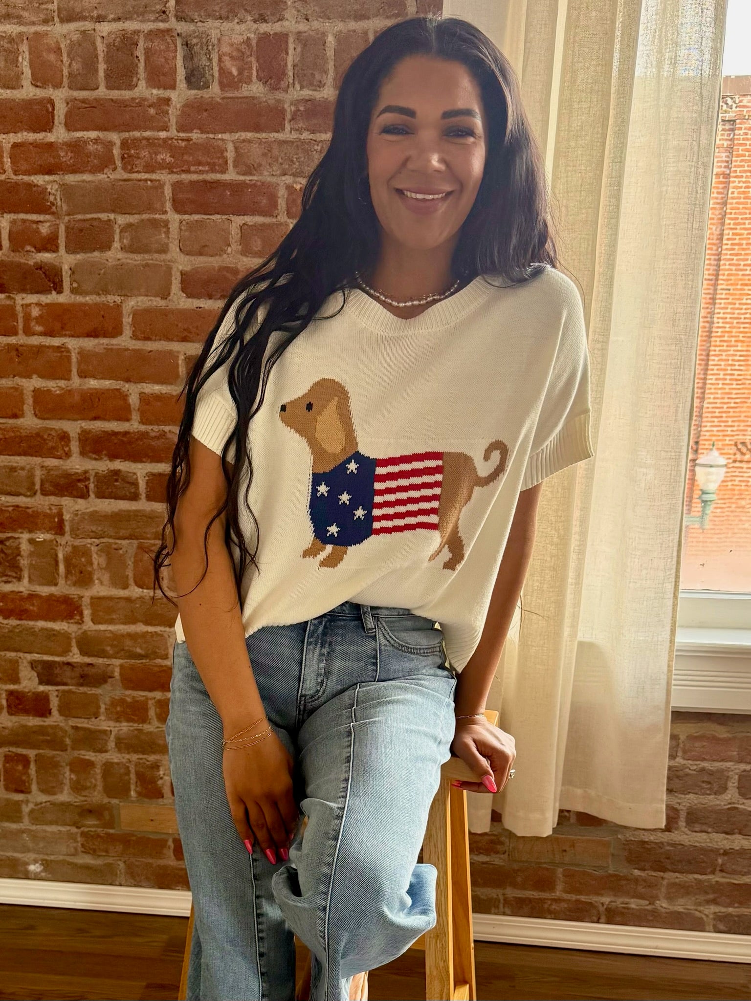 Woman wearing a white t-shirt with a dog and American flag design, standing in front of a brick wall.