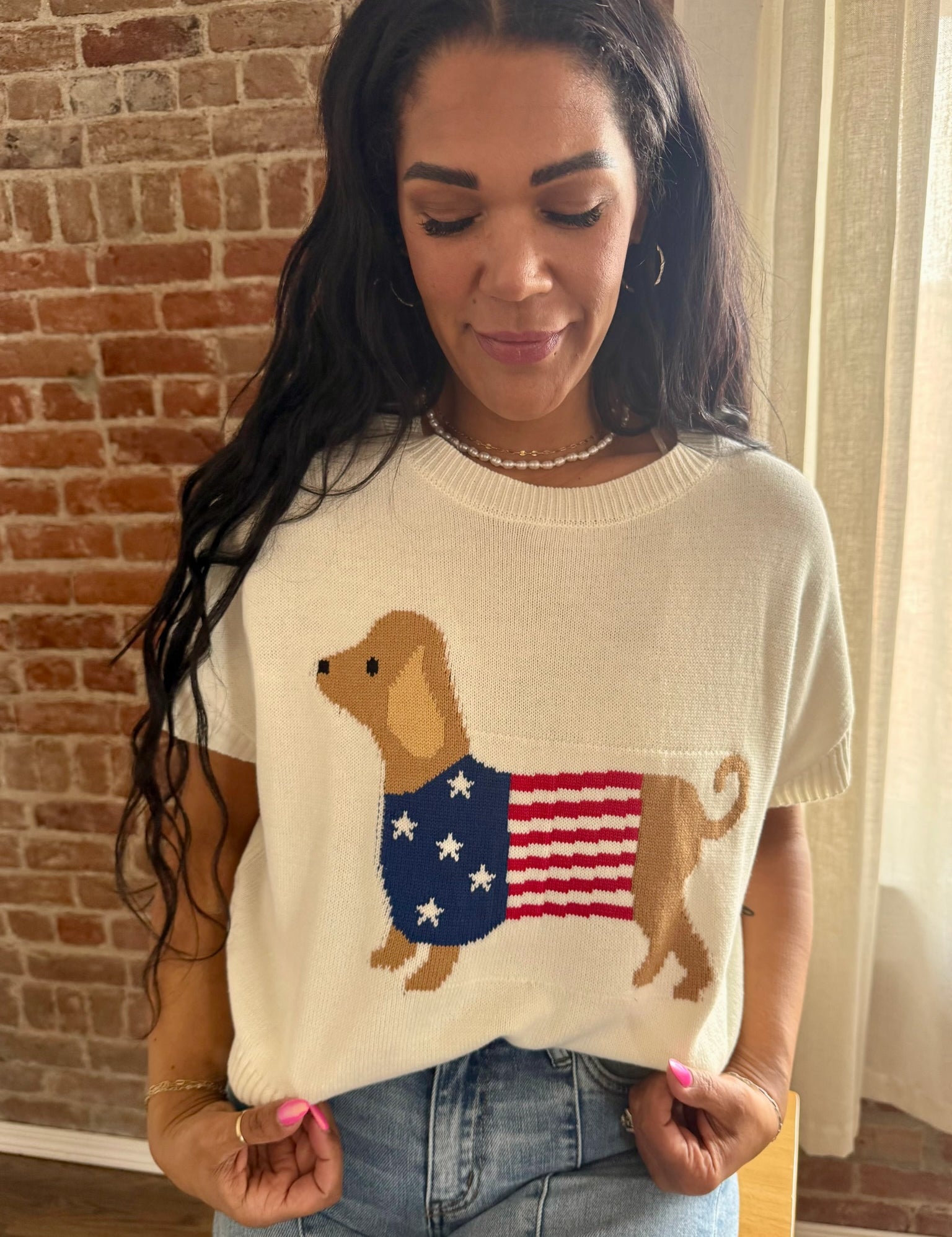 Woman wearing a sweater with a dog and American flag design, standing against a brick wall.