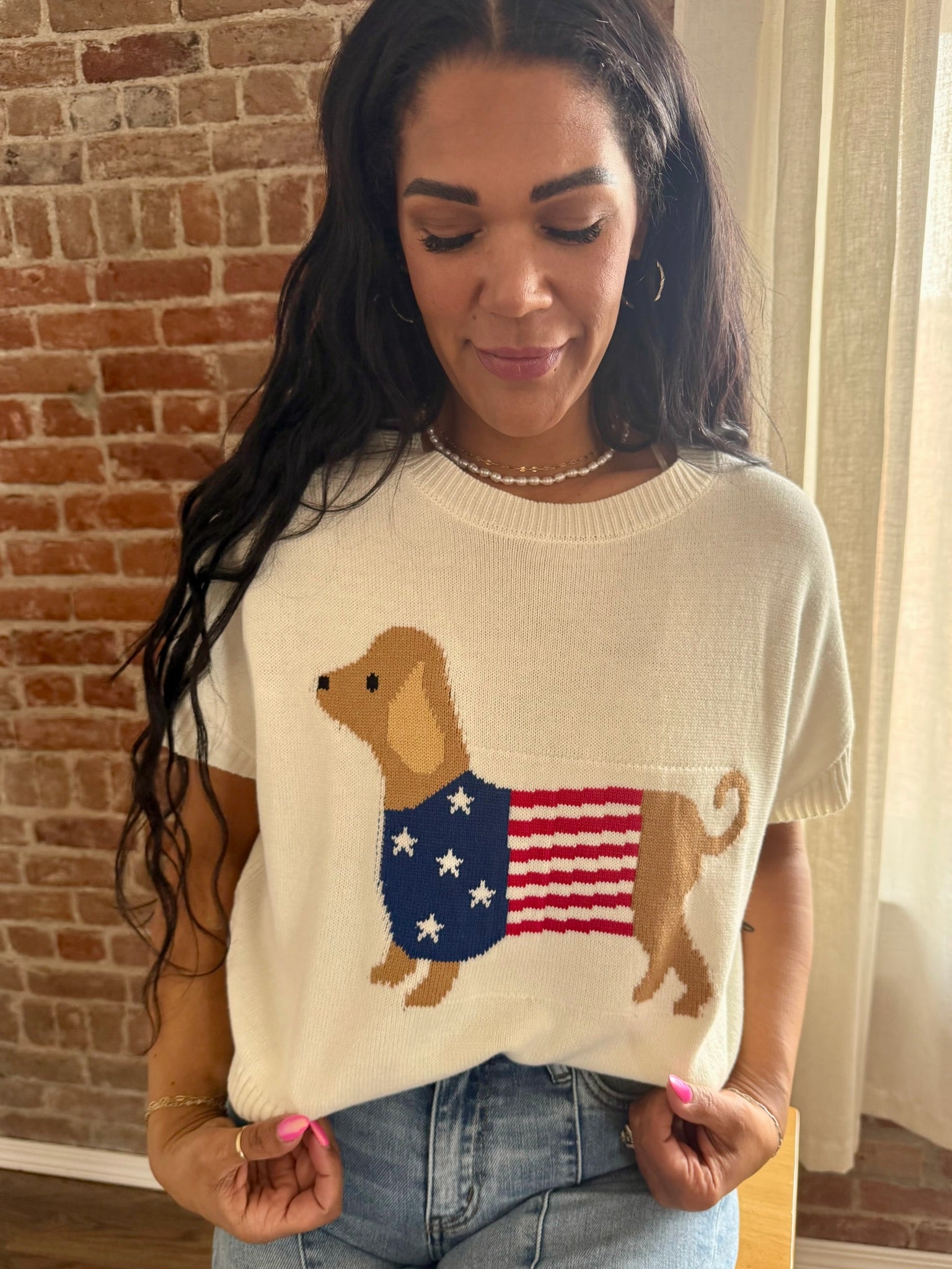 Woman wearing a sweater with a dog and American flag design, standing against a brick wall.