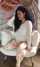 Woman sitting in a wicker chair wearing a white lace top and floral pants.