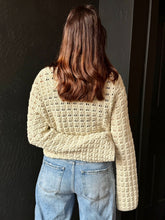 Person wearing a beige crochet sweater and blue jeans against a dark background