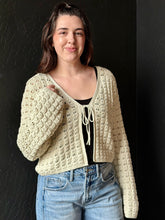 Woman wearing a beige crochet cardigan over a black top with blue jeans against a dark background