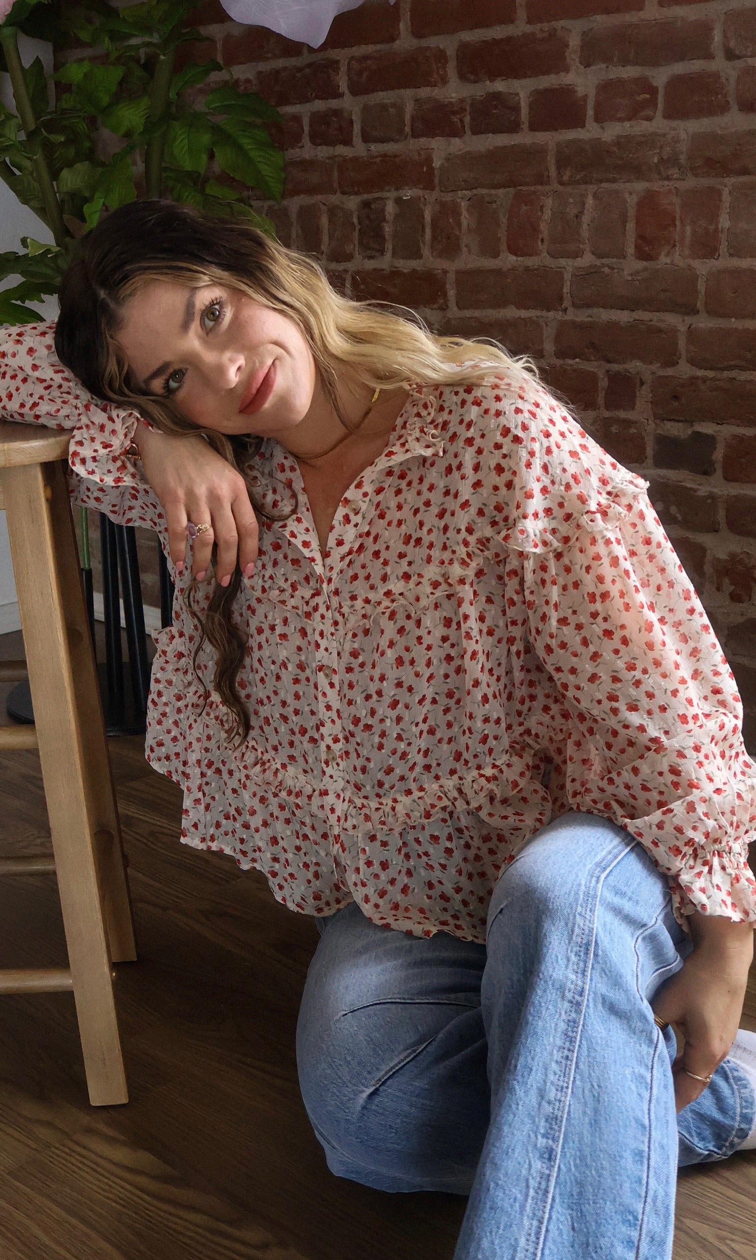 Woman in a floral blouse and jeans sitting against a brick wall.