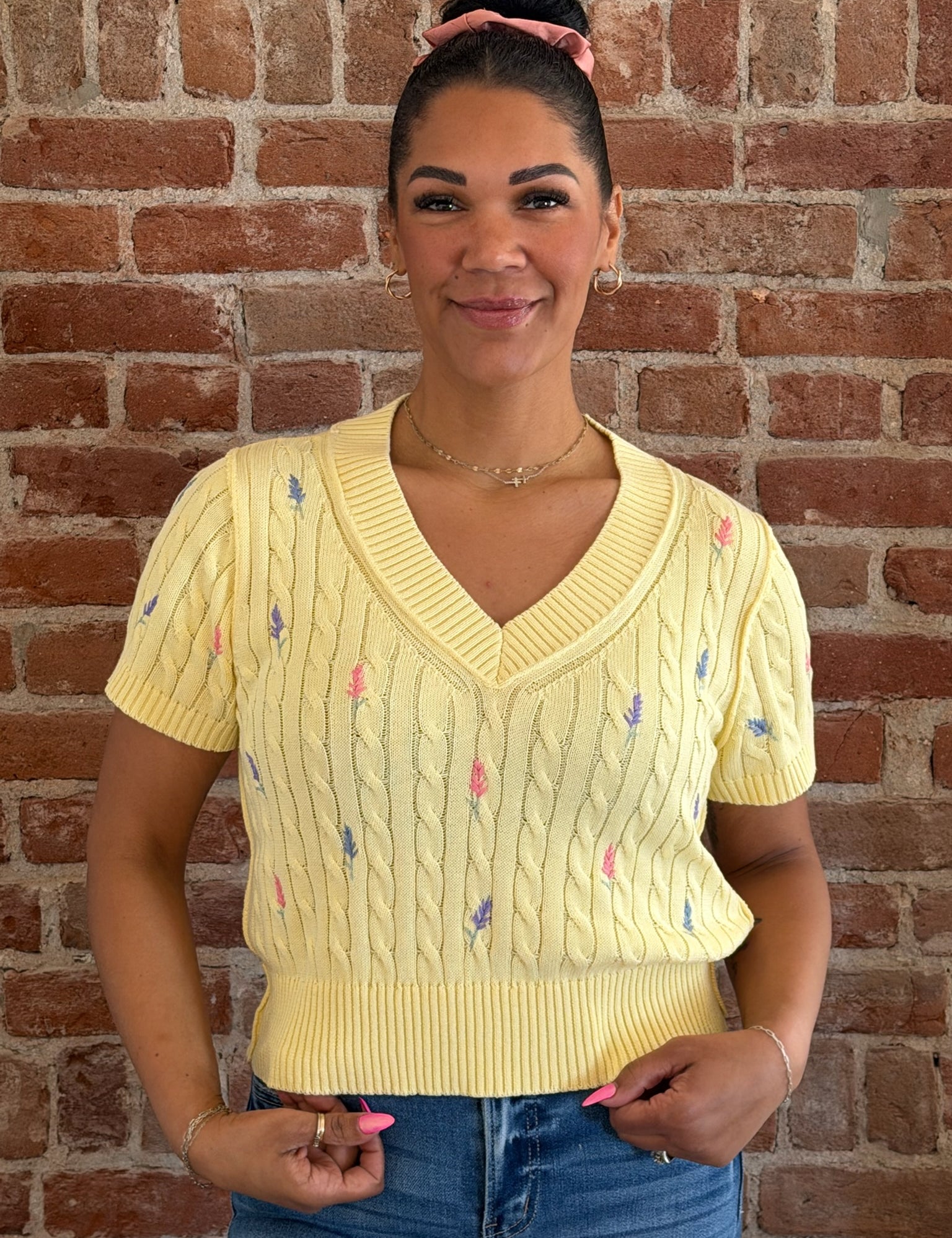 Woman wearing a yellow sweater with floral patterns against a brick wall.