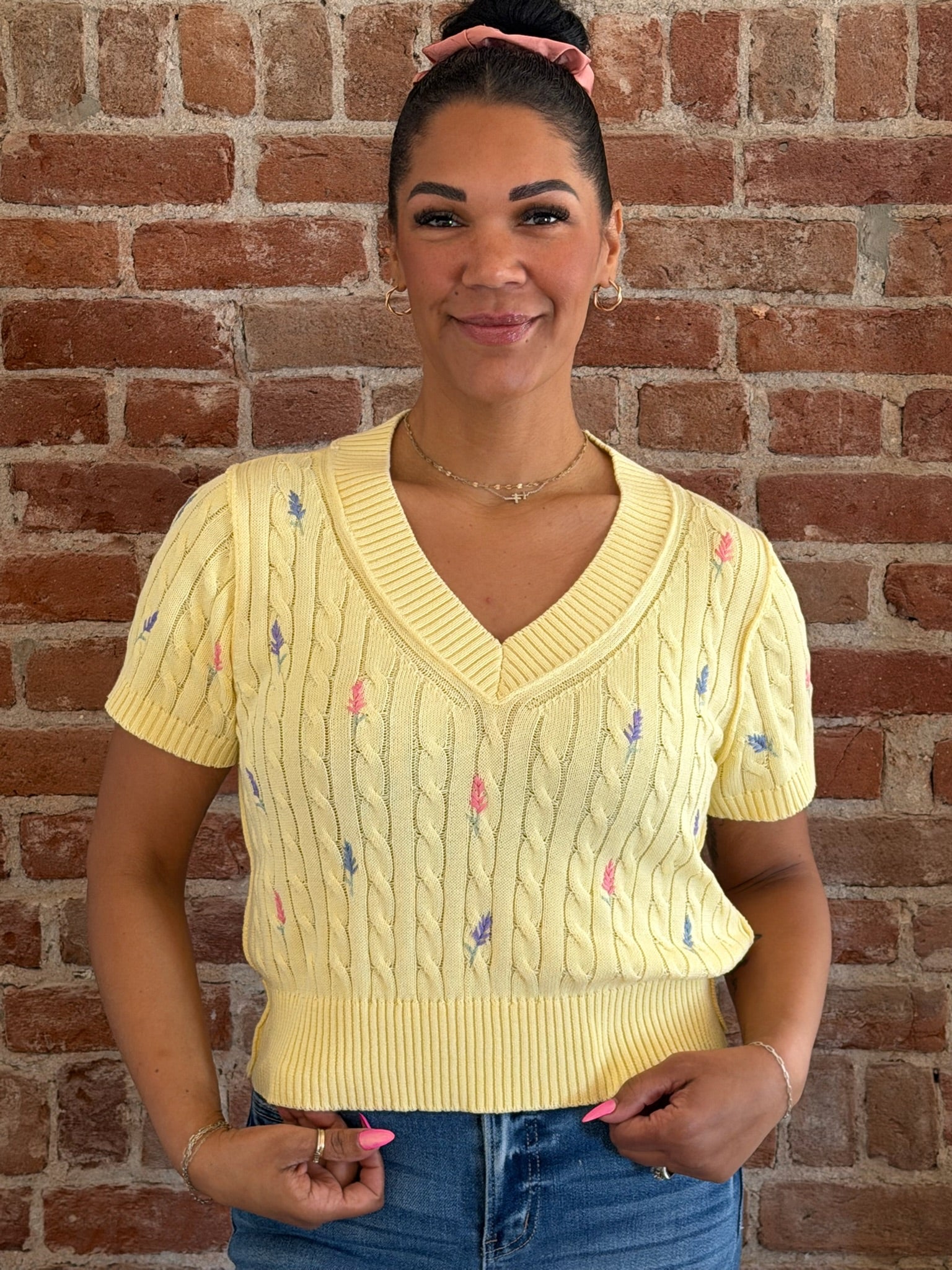 Woman wearing a yellow sweater with floral patterns against a brick wall.