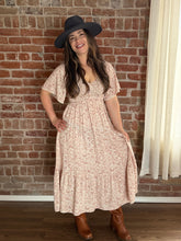 Woman wearing a floral dress and black hat against a brick wall.