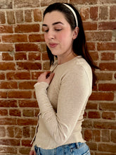 Woman wearing a beige long-sleeve top and blue jeans against a brick wall.