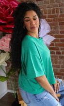 Woman wearing a green top and blue jeans sitting in front of a brick wall with flowers.