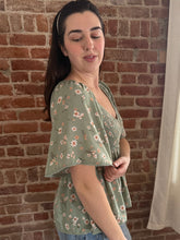 Woman wearing a green floral blouse against a brick wall.