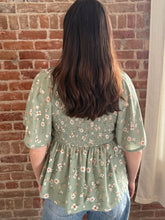 Person wearing a green floral blouse standing against a brick wall.