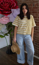 Woman in a striped shirt and jeans holding a hat, standing in front of a brick wall with large flowers.