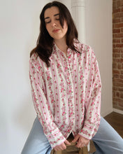 Woman wearing a pink floral shirt sitting on a stool against a white wall with a brick column.