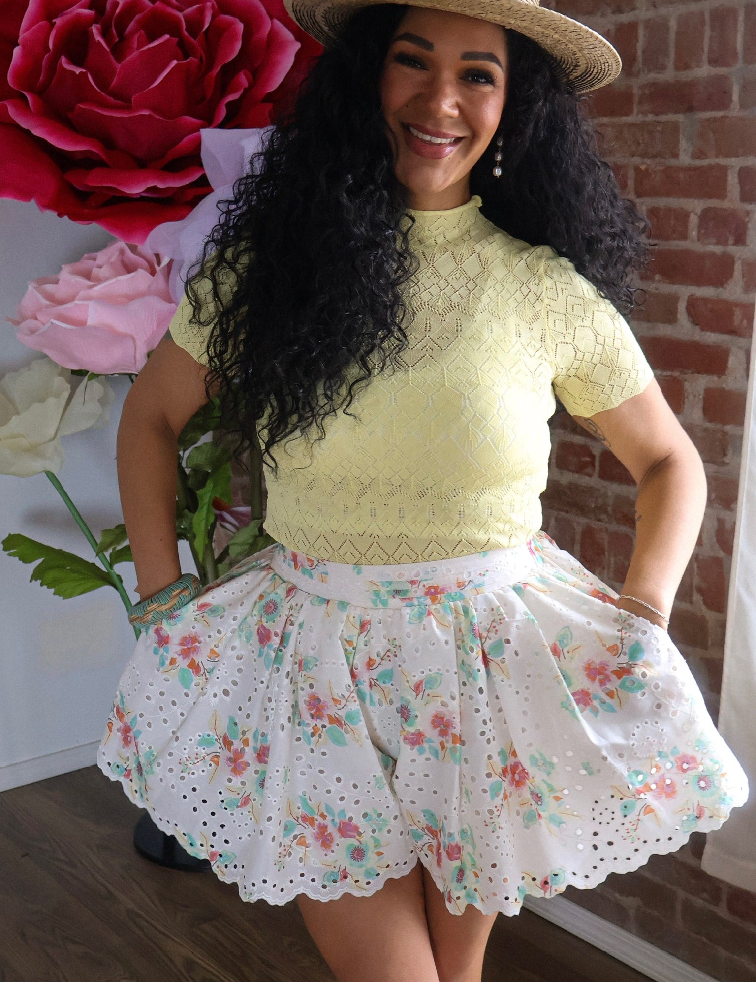 Woman wearing a yellow lace top and floral skirt with a straw hat, standing in front of a brick wall.