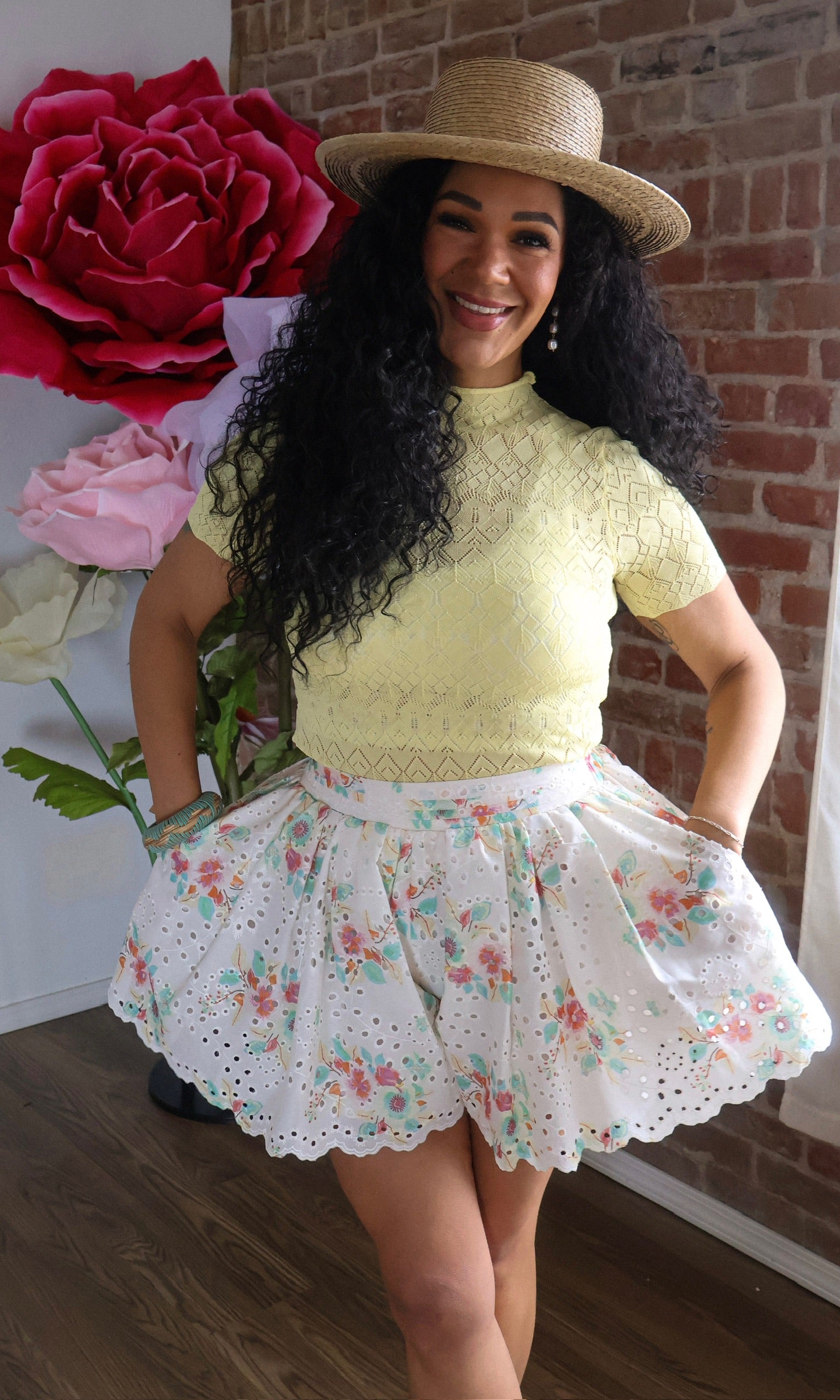 Woman wearing a yellow lace top and floral skirt with a straw hat, standing in front of a brick wall.