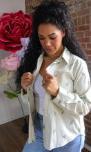 Woman wearing a light-colored striped shirt with floral patterns, standing next to large flowers indoors.