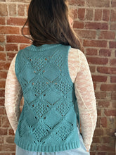 Person wearing a teal knitted vest with a brick wall background