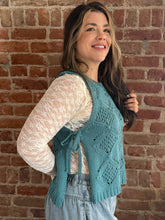 Woman wearing a teal knitted vest over a white lace top against a brick wall.