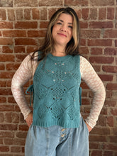 Woman wearing a teal knitted vest over a white shirt against a brick wall.