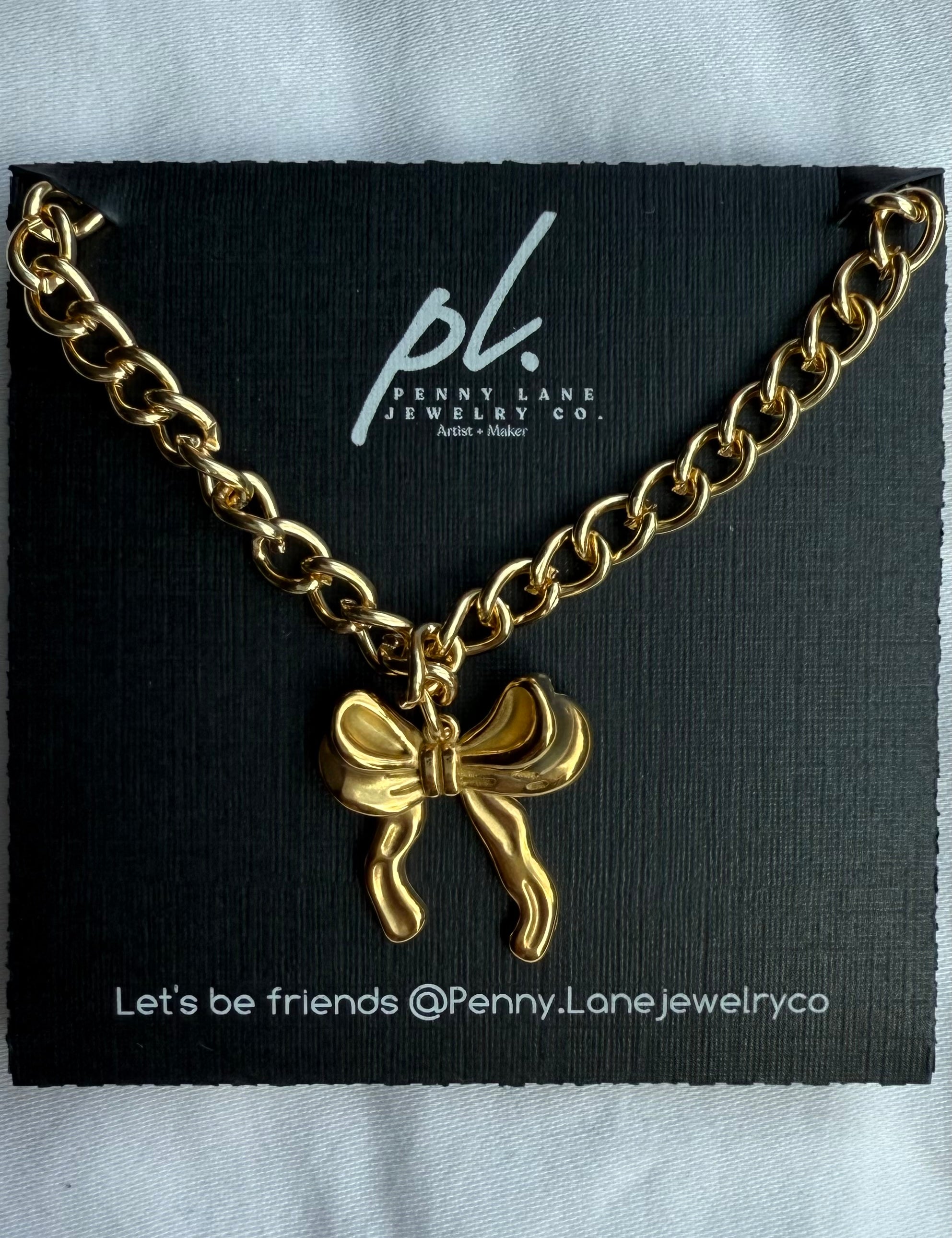 Gold chain necklace with a bow charm on a Penny Lane Jewelry Co. card.