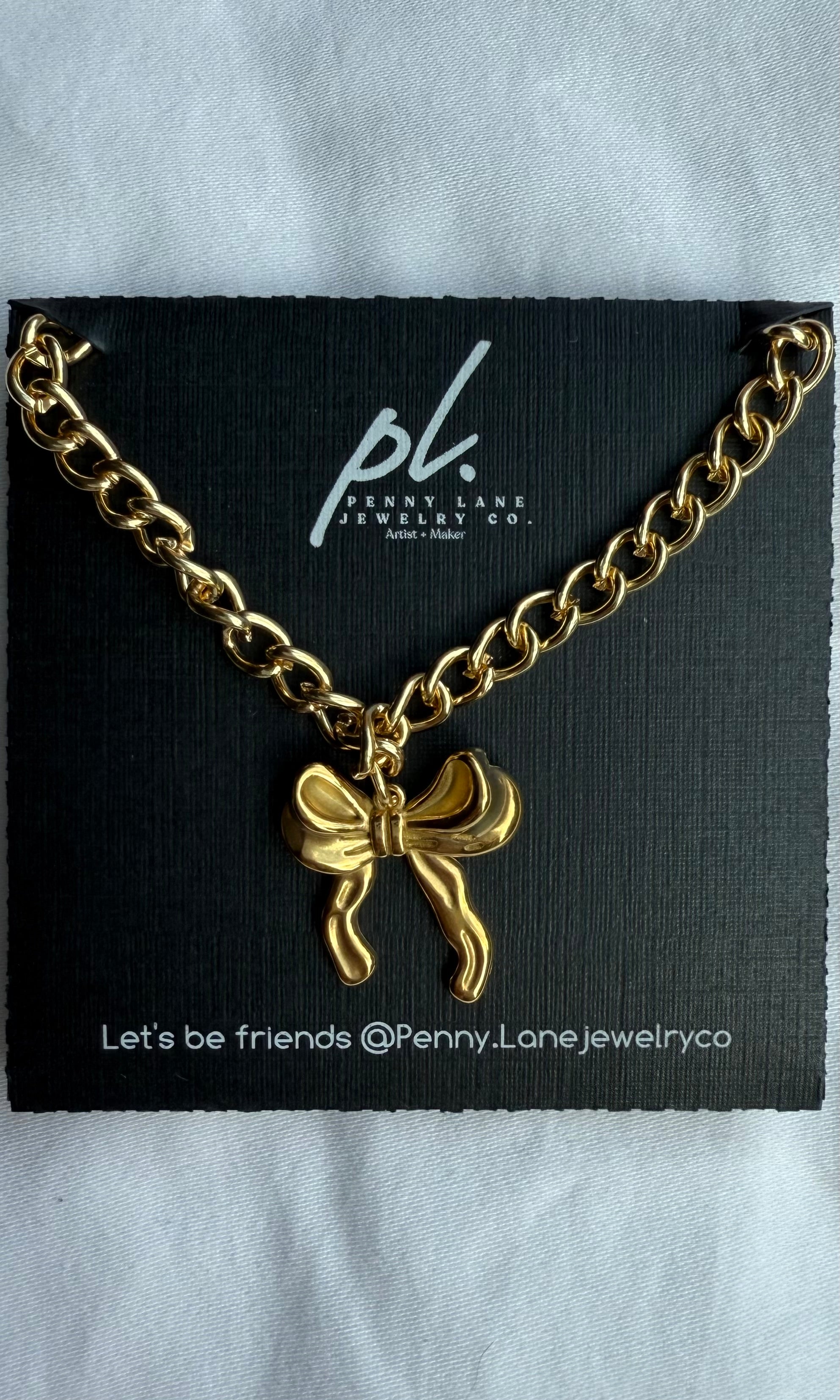 Gold chain necklace with a bow charm on a Penny Lane Jewelry Co. card.
